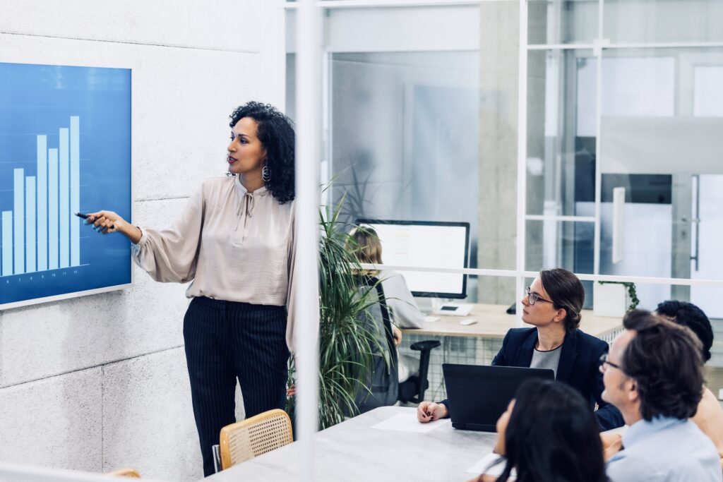 Businesswoman pointing at statistics on digital screen, giving presentation in conference room. Corporate coworkers listening to financial analytics report in office meeting