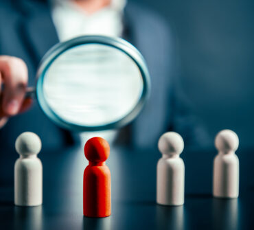 A businessperson holds a magnifying glass to a row of pawns, one of which is red in contrast with the others.